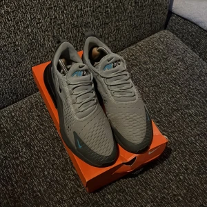 Nike Air Max 270 grå sneakers - Säljer ett par grå Nike Air Max 270 sneakers med blå detaljer och svart sula. Skorna har ventilerande mesh-ovandel och klassisk Air Max-dämpning i hälen. Perfekta för dig som vill ha både stil och komfort.