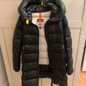Svart dunjacka från Parajumpers SHEEN - Säljer en svart lång dunjacka från Parajumpers, modell SHEEN. Jackan har hög krage, huva och dragkedja framtill. Insidan är ljus med märkets logga och detaljer. Perfekt för kalla dagar och riktigt snygg passform.