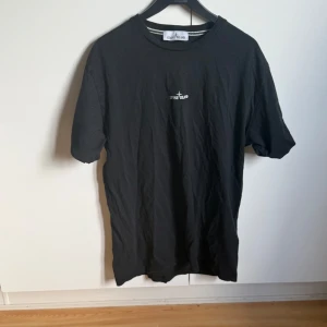 Svart t-shirt från Stone Island - Svart t-shirt från Stone Island med liten logga framtill och stor  logga på ryggen. Klassisk rund halsringning och korta ärmar. Perfekt för sommarn och vill ha en enkel men snygg look.