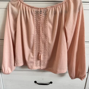 Ljusrosa offshoulder blus med spetsdetaljer - Säljer en ljusrosa offshoulder blus med långa ärmar och fina spetsdetaljer framtill. Blusen har dekorativa knytband med tofsar och ett luftigt, lätt material som ger en romantisk känsla.