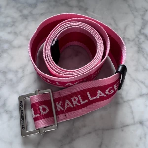 Rosa bälte från Karl Lagerfeld - Säljer ett rosa bälte från Karl Lagerfeld med tydlig logga längs hela bandet. Bältet har en silverfärgad metallspänne och är gjort i ett slitstarkt tygmaterial. Perfekt för att ge outfiten en färgklick.