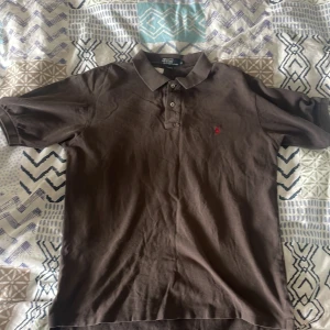 Brun/Svart pikétröja från Polo Ralph Lauren - Säljer en klassisk brun/svart pikétröja från Polo Ralph Lauren med korta ärmar och krage. Tröjan har knappar vid halsen och den ikoniska röda loggan broderad på bröstet. Perfekt för en stilren och avslappnad look.