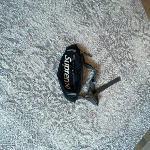 Svart axelväska från Supreme - Säljer en svart axelväska från Supreme med stort vitt logotryck på framsidan. Väskan har justerbart band och dragkedja upptill. Perfekt för att bära med sig det viktigaste.