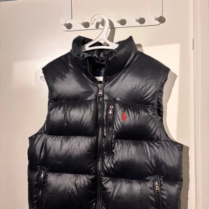 Svart dunväst från Polo Ralph Lauren - Snygg svart dunväst från Polo Ralph Lauren med dragkedja framtill och två fickor med dragkedja. Klassisk design med röd broderad logga på bröstet och hög krage. Perfekt för lager-på-lager under kyliga dagar. Nypris 2800:-