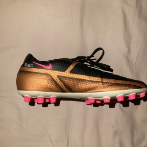 Nike Phantom GX fotbollsskor svart/guld - Säljer ett par Nike Phantom GX fotbollsskor i svart och guld med vita och rosa detaljer. Skorna har snörning, rund tå och platt sula med rosa dobbar. Perfekta för dig som vill sticka ut på planen!