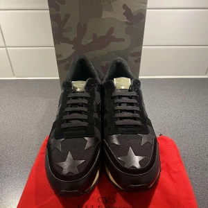 Svarta sneakers med stjärnor från Valentino Garavani - Säljer ett par svarta Valentino Garavani sneakers med stora stjärnor i grått och svart på ovandelen. Skorna har svarta snören, platt sula och detaljer i läder och textil. Kommer med originalkartong och röd dustbag.