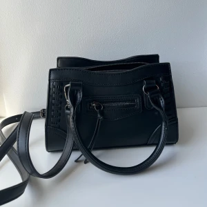 Svart axelväska från Gina Tricot - Snygg svart axelväska med både handtag och lång axelrem. Väskan har dragkedja upptill, dekorativa sömmar och en liten ficka framtill. 