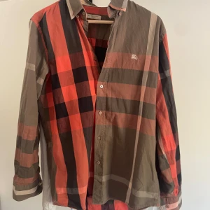 Rutig skjorta från Burberry Brit - Säljer en snygg rutig skjorta från Burberry Brit i färgerna röd, svart, beige och grå. Skjortan har klassisk krage, knappar framtill och lång ärm. Ikoniskt Burberry-mönster och liten broderad logga på bröstet.