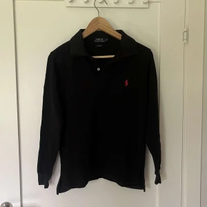 Ralph lauren piké tröja  - Klassisk svart pikétröja från Polo Ralph Lauren med lång ärm och röd broderad logga på bröstet. Tröjan har krage och knäppning framtill. Perfekt för en stilren och tidlös look.
