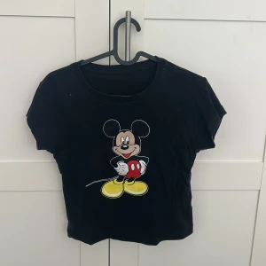 Svart croppad t-shirt med Musse Pigg-tryck - Svart croppad t-shirt med färgglatt tryck av Musse Pigg på framsidan. T-shirten har rund halsringning och korta ärmar. Den är väldigt enkel och lekfull för dem som har den stilen.