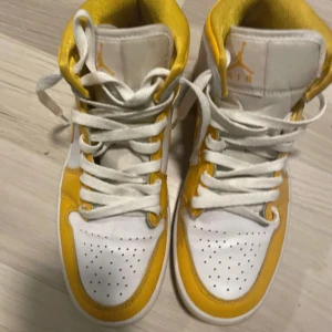 Nike Air Jordan 1 vit/gul sneakers - Nike Air Jordan 1 sneakers i vitt och gult med klassisk high-top siluett. Skorna har snörning, perforerad tå och ikoniska Jordan Wings-loggan på sidan. Tillverkade i skinn med vit bas och gula detaljer samt matchande gula yttersulor. Perfekta för dig som vill sticka ut.  Använda få gånger sparsamt använda