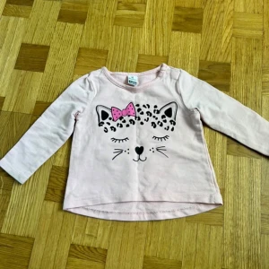 Ljusrosa långärmad tröja Bonitera - Supersöt ljusrosa långärmad tröja från Bonitera med ett gulligt kattansikte framtill, leopardmönster och en rosa prickig rosett. Tillverkad i mjuk bomull, perfekt för små barn. Rund halsringning och bekväm passform.