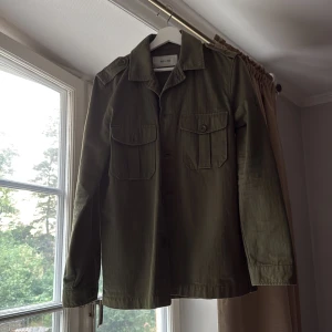 Olivgrön overshirt från Whyred - Snygg olivgrön overshirt från Whyred med klassisk krage, axelklaffar och två stora bröstfickor med knappar. Jackan är i bomull och har en rak passform med långa ärmar. Perfekt att slänga över en hoodie eller t-shirt för en chill look.