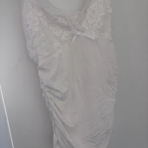 Vit linne med rosett - Supersöt vit linne med spets upptill och liten rosett vid bysten. Linnet har v-ringning, blommigt spetsmönster och är ärmlös. Perfekt för dig som gillar romantisk och feminin stil.