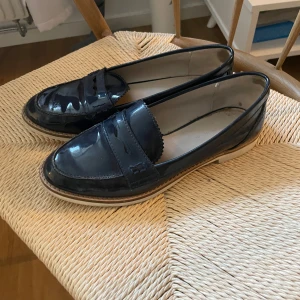 Mörkblå lackloafers med platt sula - Snygga mörkblå loafers i lackat skinn med klassisk pennyloafers-design och platt sula. Skorna har rund tå och ljus sula som kontrast. Perfekta för dig som vill ha en stilren och trendig look.