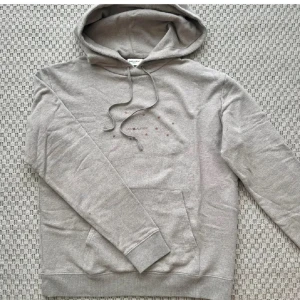 Grå hoodie från Saint Laurent - Snygg grå hoodie från Saint Laurent med huva och snörning. Tryck med små stjärnor och logga på bröstet. Mjuk bomullskänsla och klassisk känguruficka framtill. Perfekt för en chill och stilren look. Topp skick, passar som en Medium, kan tolkas någorlunda beige i verkligheten! 
