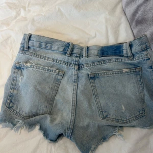 Ljusa jeansshorts från Zara - Säljer ett par ljusblå jeansshorts från Zara med råa, fransiga kanter och slitna detaljer. Shortsen har hög midja, klassiska fem fickor och knappar framtill. Perfekta för en chill sommarlook.