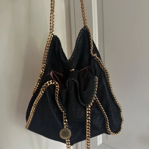 Svart Stella McCartney axelväska med kedja - Säljer min svarta axelväska från Stella McCartney med snygga guldfärgade kedjor och rund logotypbricka. Har tyvärr inget kvitto då jag köpte den av en tjej för ett halv år sedan. Har andvänt den någon gång men är i mycket bra skick. 