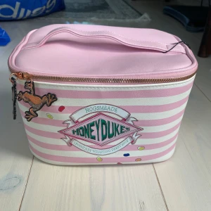 Honeydukes necessär Harry Potter - Supergullig rosa och vit randig väska från Primark x Harry Potter, inspirerad av Honeydukes. Väskan har dragkedja med chokladgroda-detaljer och ett handtag upptill. Perfekt för att förvara smink eller småprylar. Materialet är syntet och designen är riktigt söt och unik.