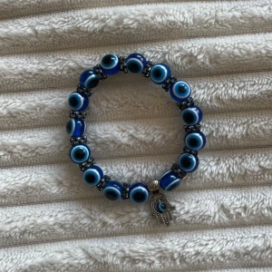Blått armband med evil eye och hamsa - Coolt armband med blå och vita evil eye-pärlor och en silverfärgad hamsa-hand som berlock. Pärlorna är runda och sitter på elastisk tråd, perfekt för dig som gillar unika smycken med symbolik.