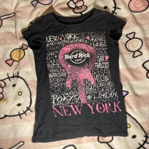 Svart Hard Rock Cafe t-shirt New York - Svart t-shirt från Hard Rock Cafe med coolt New York-tryck och rosa glitterdetaljer framtill. Kortärmad modell i 100% bomull med figurnära passform. Perfekt för dig som gillar statement-plagg och musikvibes.