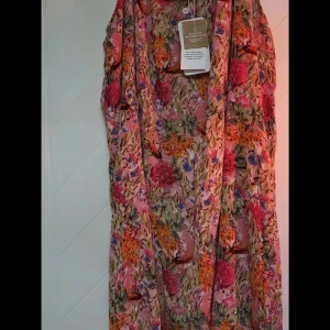 Blommig klänning från Zhenzi - Färgstark klänning från Zhenzi med blommigt mönster i rosa, orange, blått och grönt. Klänningen är ärmlös och tillverkad i hållbar viskos, perfekt för sommarens varma dagar. Snygg och flowy passform som ger en bohemisk vibe.