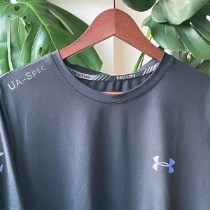 Under Armour t-shirt - Svart t-shirt från Under Armour med reflekterande logga på bröstet och tryck på axeln. Tillverkad i ett lätt och snabbtorkande syntetmaterial, perfekt för löpning eller gym. Klassisk passform med rund hals och korta ärmar.