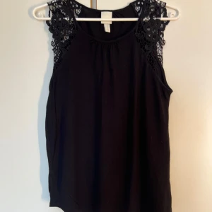 Svart ärmlös topp med spets H&M XS - Svart ärmlös topp från H&M i storlek XS. Toppen har snygga spetsdetaljer med blommönster på axlarna och en rund halsringning. Perfekt för dig som gillar en stilren look med lite extra detaljer.