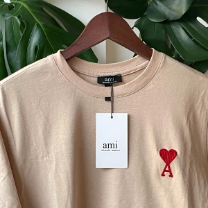 Ami t-shirt  - Storlek: S men passar perfekt till M. Snygg beige t-shirt från Ami med röd broderad hjärtdetalj och bokstaven A på bröstet. Klassisk rund halsringning och korta ärmar. Tillverkad i mjuk bomull som känns skön mot huden. Perfekt för en clean och trendig look.