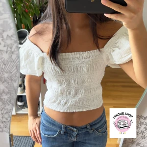Vit offshoulder smockad topp - Superfin vit topp med smockad kropp och puffiga offshoulder-ärmar. Croppad modell med fyrkantig ringning och elastisk passform som sitter snyggt. Perfekt till jeans eller kjol för en fräsch look.