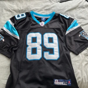 Carolina Panthers Smith #89 NFL-tröja - Säljer en svart Carolina Panthers NFL-tröja från Reebok med nummer 89 och namnet Smith på ryggen. Tröjan har blå och silverfärgade detaljer, stora tryckta siffror och Panthers-logga på ärmarna. Tillverkad i ventilerande polyester med korta ärmar och V-ringning.