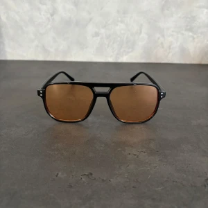 Svarta pilotbågar med bruna glas - Snygga solglasögon med klassisk pilotform och svarta bågar i plast. Glasen är bruna och ger en cool retrovibe. Perfekta för dig som vill sticka ut med en tidlös stil. Passar både till city och strandhäng.