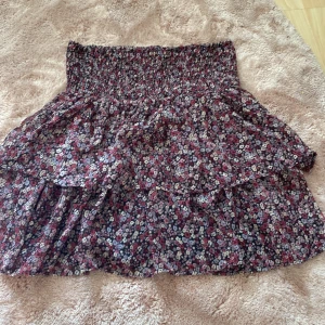  Lila Volangkjol från Kappahl - Supersöt kort volangkjol från Kappahl med blommigt mönster i rött, lila, vitt och svart. Kjolen har smock i midjan och flera lager volanger som ger en flowy känsla. Perfekt för dig som gillar romantisk och färgglad stil!