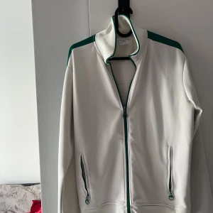 Vit och grön vindjacka från Zara - Sportig vit vindjacka från Zara med gröna detaljer längs axlar och dragkedjor. Jackan har hög krage, hel dragkedja framtill och två fickor med dragkedja. Perfekt för en chill och avslappnad stil.