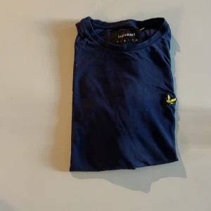 Marinblå t-shirt från Lyle & Scott - Snygg marinblå t-shirt från Lyle & Scott med klassisk gul logga broderad på bröstet. T-shirten är i mjuk bomull och har en normal passform med korta ärmar och rund hals. Perfekt för en clean och stilren look. Herr strl 