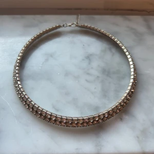Glittrigt silverhalsband med stenar - Festligt halsband i silverfärg med två rader glittrande stenar som ger en lyxig känsla. Halsbandet har en rund, stel form och stängs med en kedja och hake. 