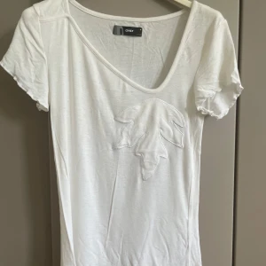 Vit t-shirt med broderad detalj från ONLY - Vit t-shirt från ONLY med kort ärm och v-ringning. T-shirten har en broderad bladformad detalj framtill och är gjord i mjuk bomull. Enkel och clean stil som passar perfekt till jeans eller shorts.