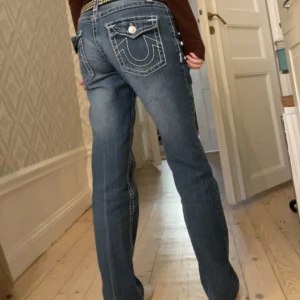 True Religion blå bootcut jeans - Säljer ett par blå jeans från True Religion med ikoniska bakfickor och kontrastsömmar. Jeansen har bootcut passform, låg midja och klassisk femficksdesign. Materialet är jeans i bomull med snygg tvätt och slitningar framtill. Perfekta för dig som gillar street och statement.