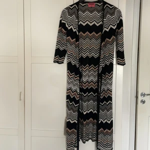 Missoni Lindex långklänning XS - Snygg långklänning från Missoni Lindex i storlek XS. Klänningen har ett ikoniskt zigzag-mönster i svart, vitt och brons, med trekvartsärm och rak passform. Materialet är stickat och har en lätt glittrig effekt som ger extra edge.