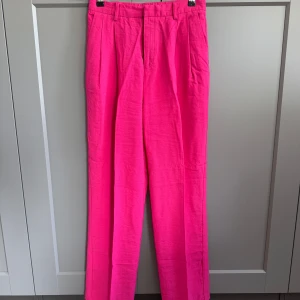 Chockrosa kostymbyxor från Lindex - Snygga kostymbyxor från Lindex i en riktigt chockrosa färg. Byxorna har raka ben, pressveck och hällor för bälte. De är midjehöga och har fickor bak. Perfekta för dig som vill sticka ut med färg!