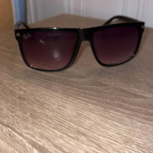 Svarta Ray-Ban solglasögon - Snygga svarta solglasögon från Ray-Ban med fyrkantig form och mörka tonade glas. Klassisk och stilren design som passar perfekt till sommaren. Diskret Ray-Ban-logga i hörnet på linsen och robust båge i plast.