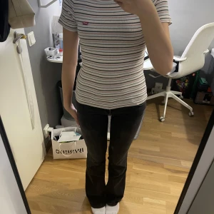 Randig ribbad topp från Levi's - Superfin ribbad topp från Levi's med smala ränder i svart, rött, blått och beige. Kortärmad och rätt tajt passform med rund halsringning. Klassisk Levi's-logga broderad vid bröstet. Perfekt till jeans eller shorts.