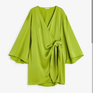 Grön omlottklänning med knyt - Snygg kort grön omlottklänning med v-ringning och vida ärmar. Klänningen har ett stort knyt i midjan som ger en cool och trendig look. Tillverkad i ett glansigt material som känns lyxigt och skönt mot huden.