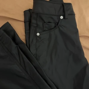 Svarta byxor i skinnimitation - Snygga svarta byxor i skinnimitation med raka ben och klassisk femficksdesign. Byxorna har silverfärgade knappar och bälteshällor, vilket ger en stilren look. Perfekta för dig som vill sticka ut med en edgy vibe.storleken är 32