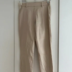 Beige byxa/slacks - Snygga beige byxor från H&M med raka ben och klassiska passpoalfickor bak. Byxorna har hällor för bälte och är tillverkade i ett mjukt bomullsmaterial. Perfekta för en clean och stilren look. Dragkedjan är på sidan