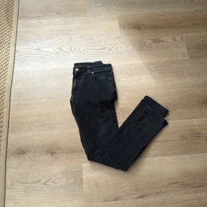 Svarta jeans från Nudie Jeans - Säljer ett par svarta jeans från Nudie Jeans. De har klassisk femficksdesign, smal passform och är tillverkade i mjuk bomullsdenim. Snygga kontrastsömmar och diskret logga på linningen. Perfekta för dig som gillar en stilren look. Har en liten liten fläck se bild 5. Storlek 30-32