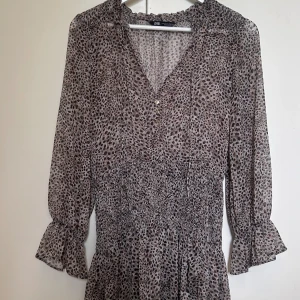 Klänning från Zara - Snygg kortklänning från Zara med leopardmönster i beige och bruna toner. Klänningen har långärmade volangärmar, smockad midja och v-ringning med knappar och knyt detalj. Tunt, luftigt material som ger en cool och trendig look.