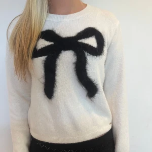 Vit stickad tröja med svart rosett - Superfin vit stickad tröja med en stor svart fluffig rosett framtill. Tröjan har rund halsringning och långa ärmar. Tröjan är knappt använd och i jättebra skick.