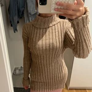 Beige ribbad polotröja - Säljer en beige ribbad polotröja med lång ärm och hög krage. Tröjan är stickad och har en loose passform med breda ribbade detaljer som ger en snygg och trendig look. Perfekt för lager-på-lager under kyliga dagar.