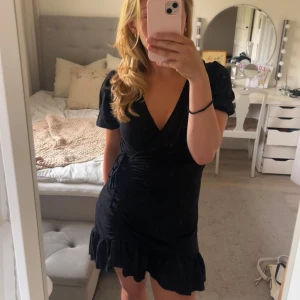 Svart omlottklänning med volang - Supersnygg svart kort omlottklänning med puffärmar, knyt i midjan och volangkant nertill. Djup v-ringning och lätt rynkade detaljer framtill ger en trendig look. Perfekt för dig som gillar stilrena och feminina plagg. Bara att skriva vid frågor. Pris går att diskutera!